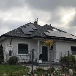 Dom jednorodzinny z szarym dachem pokrytym panelami słonecznymi; na szczycie dachu siedzi czarny ptak.