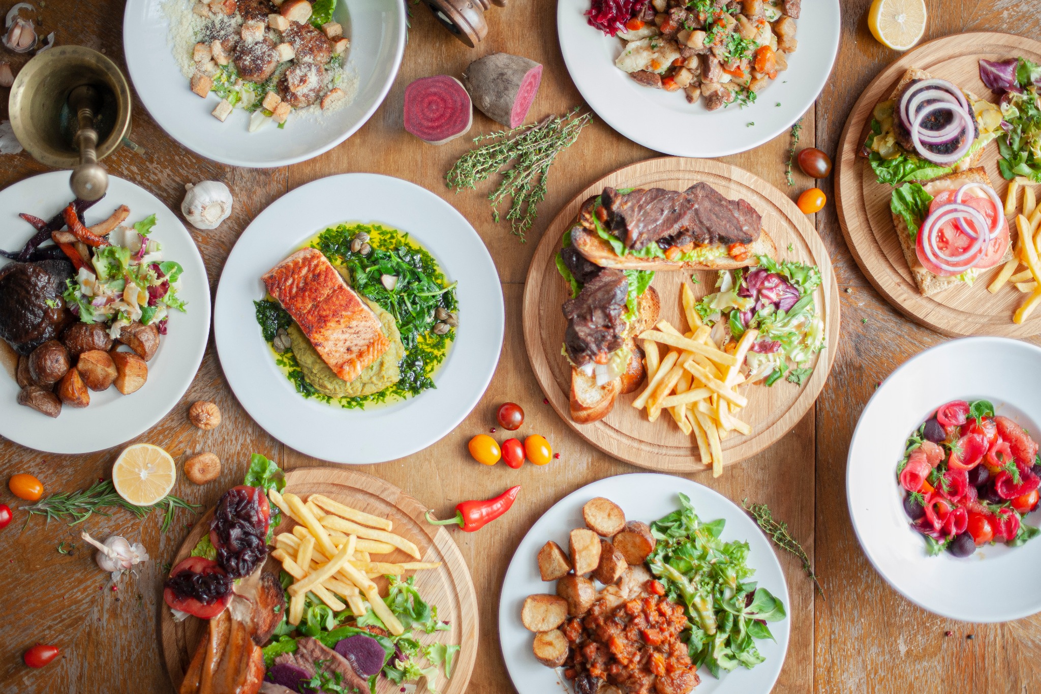 Widok z góry na drewniany stół zastawiony różnorodnymi daniami: sałatki, ryba, mięso, frytki, warzywa, udekorowane ziołami i przyprawami, podane na białych talerzach i drewnianych deskach.