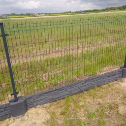 Artblok - Nowoczesne ogrodzenie panelowe z podmurówką imitującą kamień, na tle zielonego pola i pochmurnego nieba. Widoczne słupki i siatka ogrodzeniowa.