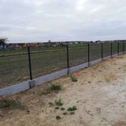 Artblok - Długi, panelowy płot z podmurówką z betonu, otoczony polem i drzewami w tle. Widok z perspektywy ziemi, pochmurne niebo.