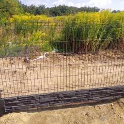 Artblok - Nowoczesne ogrodzenie panelowe z podmurówką imitującą kamień, na tle zieleni. Metalowe słupki i siatka tworzą solidną konstrukcję. Widok z bliska.