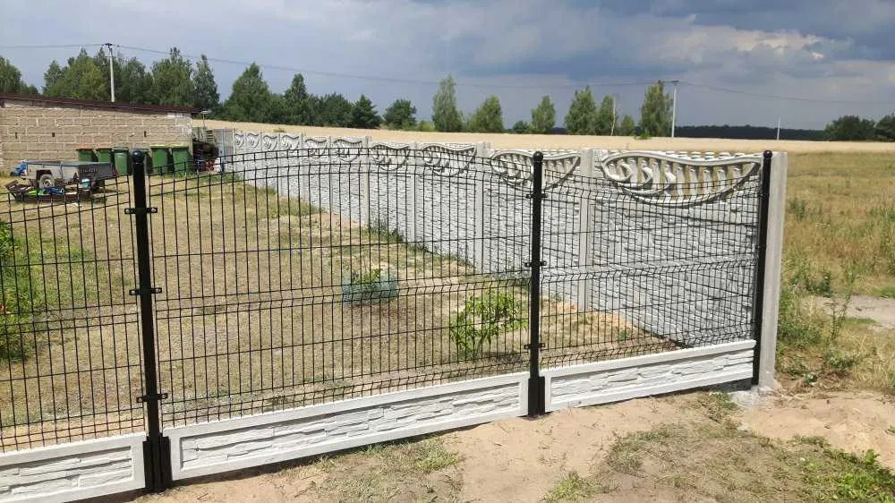 Nowoczesne ogrodzenie panelowe z podmurówką imitującą kamień, czarne słupki, widok na pole i budynek gospodarczy w tle, pochmurne niebo.