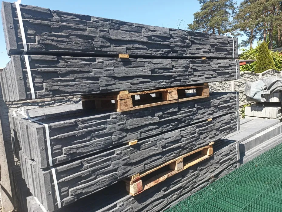 Szare, betonowe panele imitujące kamień, ułożone na paletach i zabezpieczone taśmą. W tle widoczne drzewa i fragment ogrodzenia.