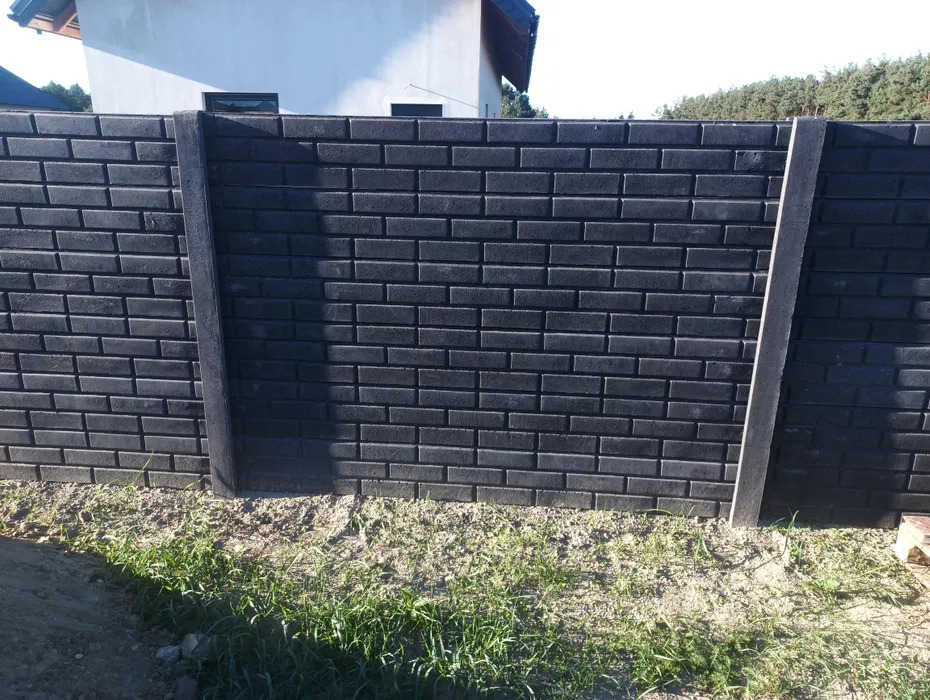 Nowoczesne ogrodzenie betonowe w Wieluniu. Ciemnoszare bloczki imitujące cegłę, z jasnymi, betonowymi słupkami. Widok z poziomu gruntu, trawa u podstawy.