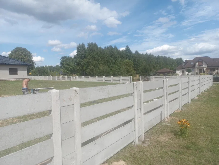 Długi, betonowy płot panelowy z przerwami, ciągnący się wzdłuż posesji. W tle domy, zieleń i błękitne niebo z chmurami. Mężczyzna pracuje przy płocie w oddali.