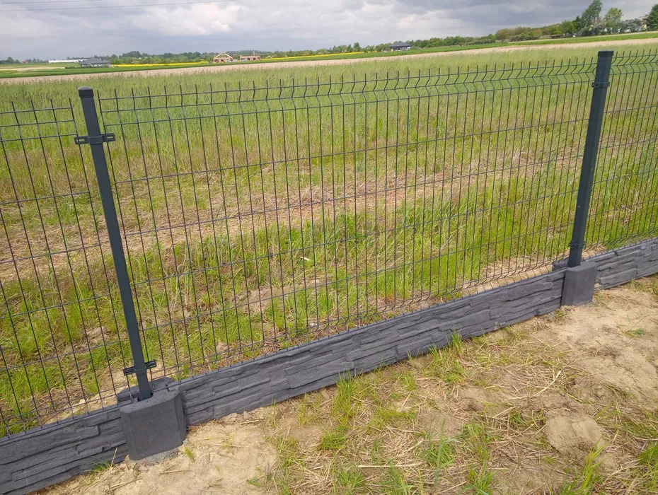 Nowoczesne ogrodzenie panelowe z podmurówką imitującą kamień, na tle zielonego pola i pochmurnego nieba. Widoczne słupki i siatka ogrodzeniowa.