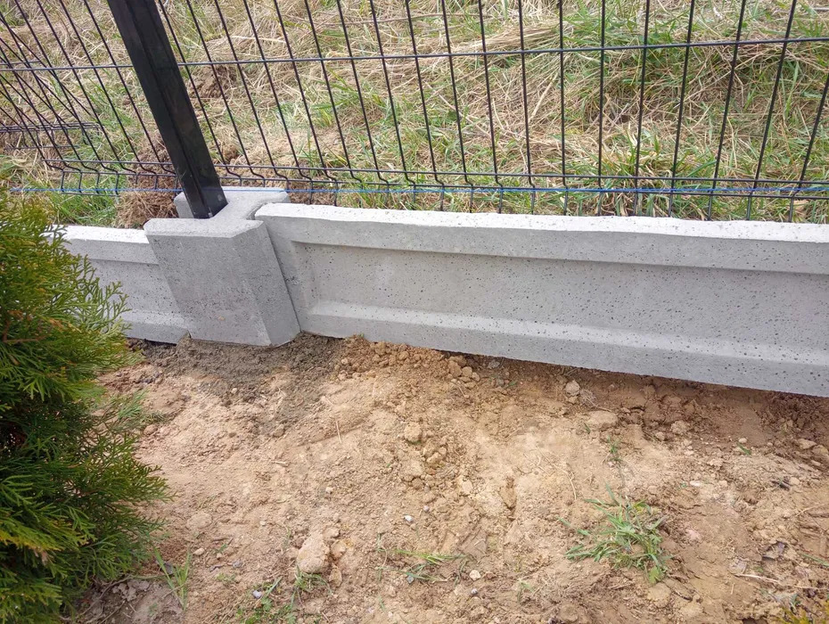 Fragment ogrodzenia betonowego z metalową siatką i słupkiem, widoczne zieleń i ziemia. Szara kolorystyka betonu i metalu.