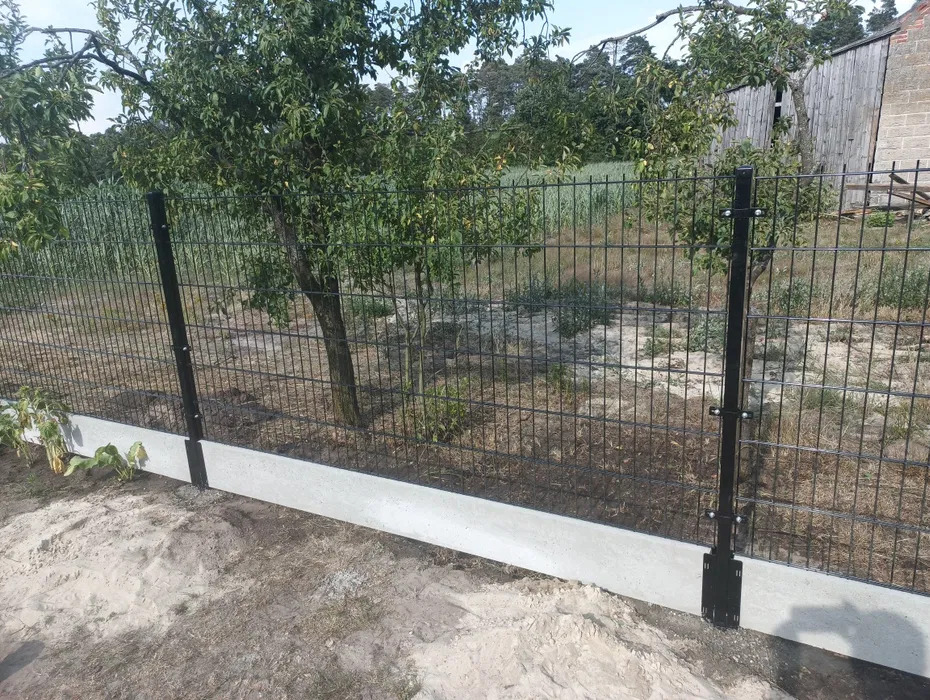 Nowoczesne ogrodzenie panelowe z podmurówką betonową, czarne słupki i siatka na tle zieleni ogrodu, widoczne elementy montażowe.