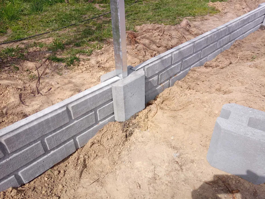 Fragment ogrodzenia betonowego z imitacją cegły, z metalowym słupkiem, na piaszczystym podłożu. Widoczny element do dalszej budowy ogrodzenia.