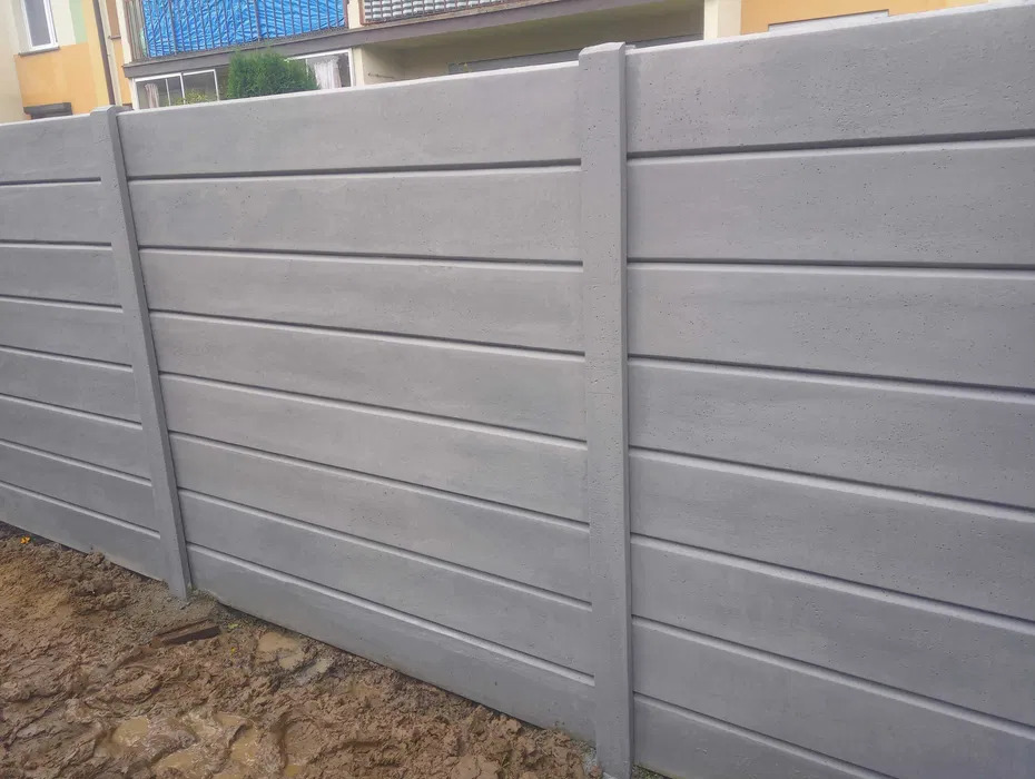 Szary, prefabrykowany płot betonowy z poziomymi panelami imitującymi deski, widoczny z bliska, z fragmentem budynku w tle i błotnistym podłożem.