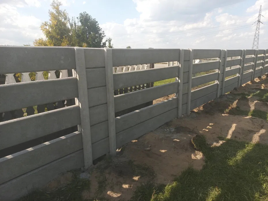 Nowoczesne ogrodzenie betonowe z poziomymi panelami, widok z bliska na tle zieleni i błękitnego nieba z chmurami. Słupy ogrodzeniowe z betonu.