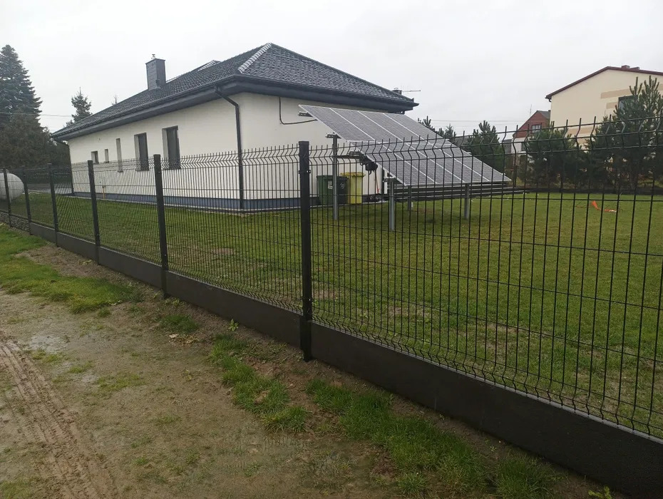 Nowoczesne ogrodzenie panelowe z podmurówką w kolorze antracytowym, na tle domu z panelami słonecznymi i zielonego trawnika. Widok z bliska, pochmurny dzień.