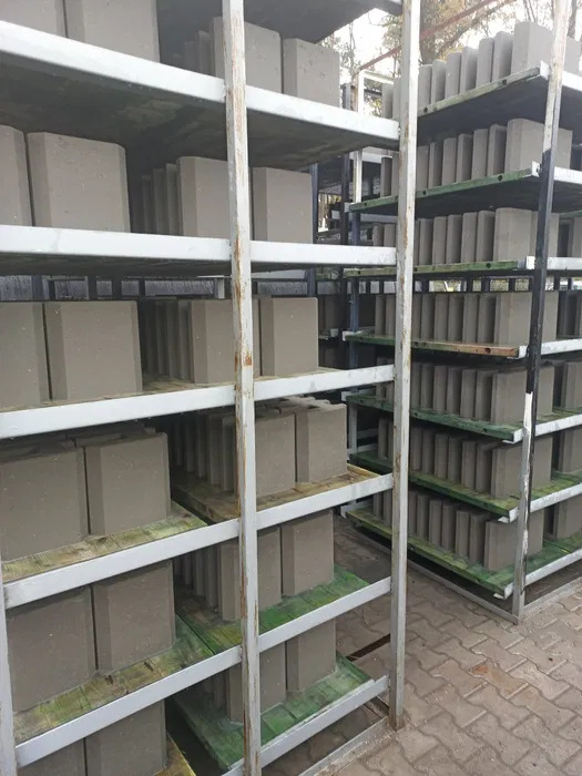 Szare bloczki betonowe ułożone na metalowych regałach w magazynie, widoczne ślady wilgoci na półkach, w tle zarys drzew.