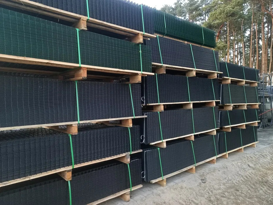 Stos palet z panelami ogrodzeniowymi w odcieniach zieleni i grafitu, zabezpieczonych zielonymi taśmami, na tle drzew. Widok z bliska, ukazujący strukturę paneli.