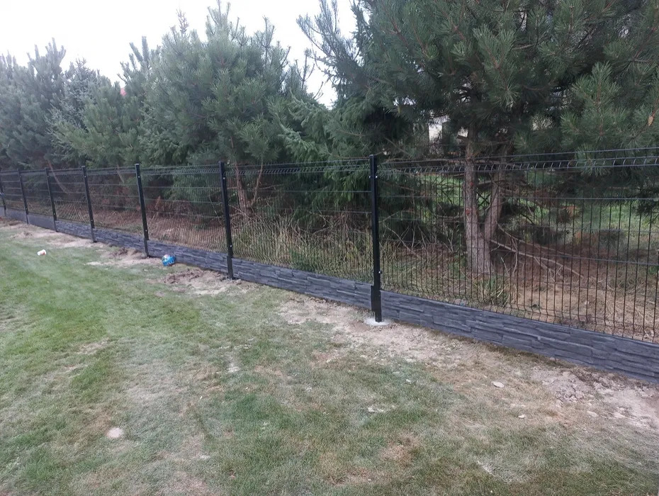 Nowoczesne ogrodzenie panelowe z podmurówką imitującą kamień, otoczone zielenią. Widoczna piłka i zabawka na trawie.