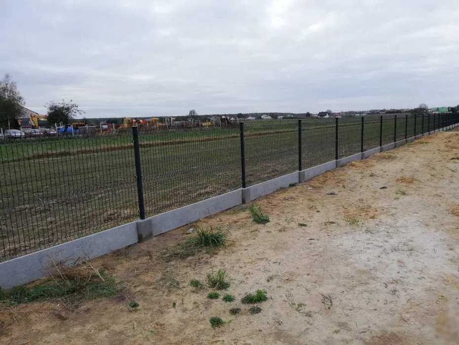 Długi, panelowy płot z podmurówką z betonu, otoczony polem i drzewami w tle. Widok z perspektywy ziemi, pochmurne niebo.