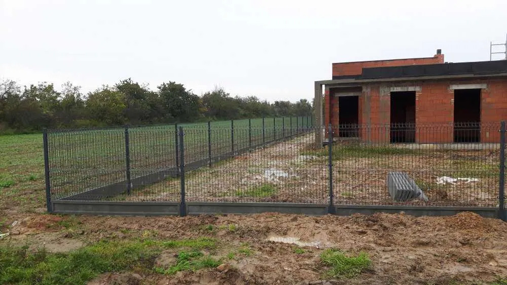 Nowoczesne ogrodzenie panelowe z podmurówką na nierównym terenie, w tle surowy budynek w trakcie budowy i zielone pole. Szara kolorystyka, pochmurny dzień.