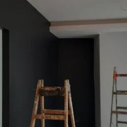 Wnętrze w trakcie malowania: ściana pomalowana na grafitowo, druga na biało, dwie drewniane drabiny malarskie w pomieszczeniu, widok na zewnątrz przez okno balkonowe.