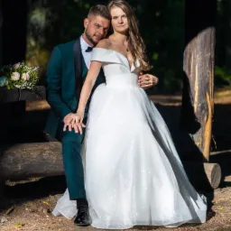 Para młoda: panna młoda w białej, błyszczącej sukni z odkrytymi ramionami i pan młody w ciemnozielonym smokingu z czarnymi klapami, siedzą na drewnianej ławce w lesie, trzymając się za ręce; bukiet...