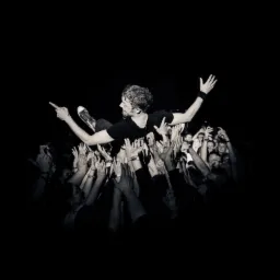 Czarno-białe zdjęcie z koncertu: wokalista na rękach publiczności, wskazujący palcem w lewo, otoczony uniesionymi rękami fanów w ciemnym otoczeniu.