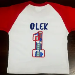 Biała koszulka z czerwonymi rękawami raglan, z niebieskim napisem 'OLEK' i cyfrą '1' w obrysie z motywem fabryki lub miasta.