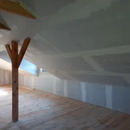 Poddasze po montażu płyt karton-gips na skosach, widoczne drewniane słupy konstrukcyjne i surowa drewniana podłoga, ściany przygotowane do dalszych prac wykończeniowych.