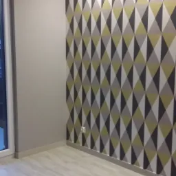 Pokój po remoncie: ściana z geometryczną tapetą w odcieniach szarości, bieli i żółci, jasna podłoga imitująca drewno, widoczne gniazdko elektryczne przy podłodze.