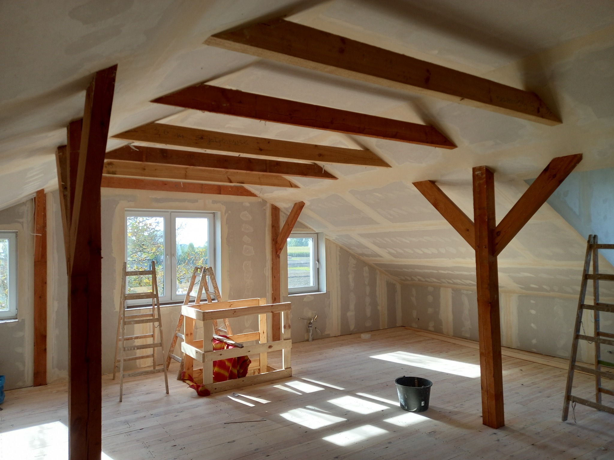 Poddasze w trakcie remontu: drewniane belki konstrukcyjne, częściowo położone płyty gipsowo-kartonowe, drabiny i rusztowanie w jasnym, słonecznym wnętrzu.