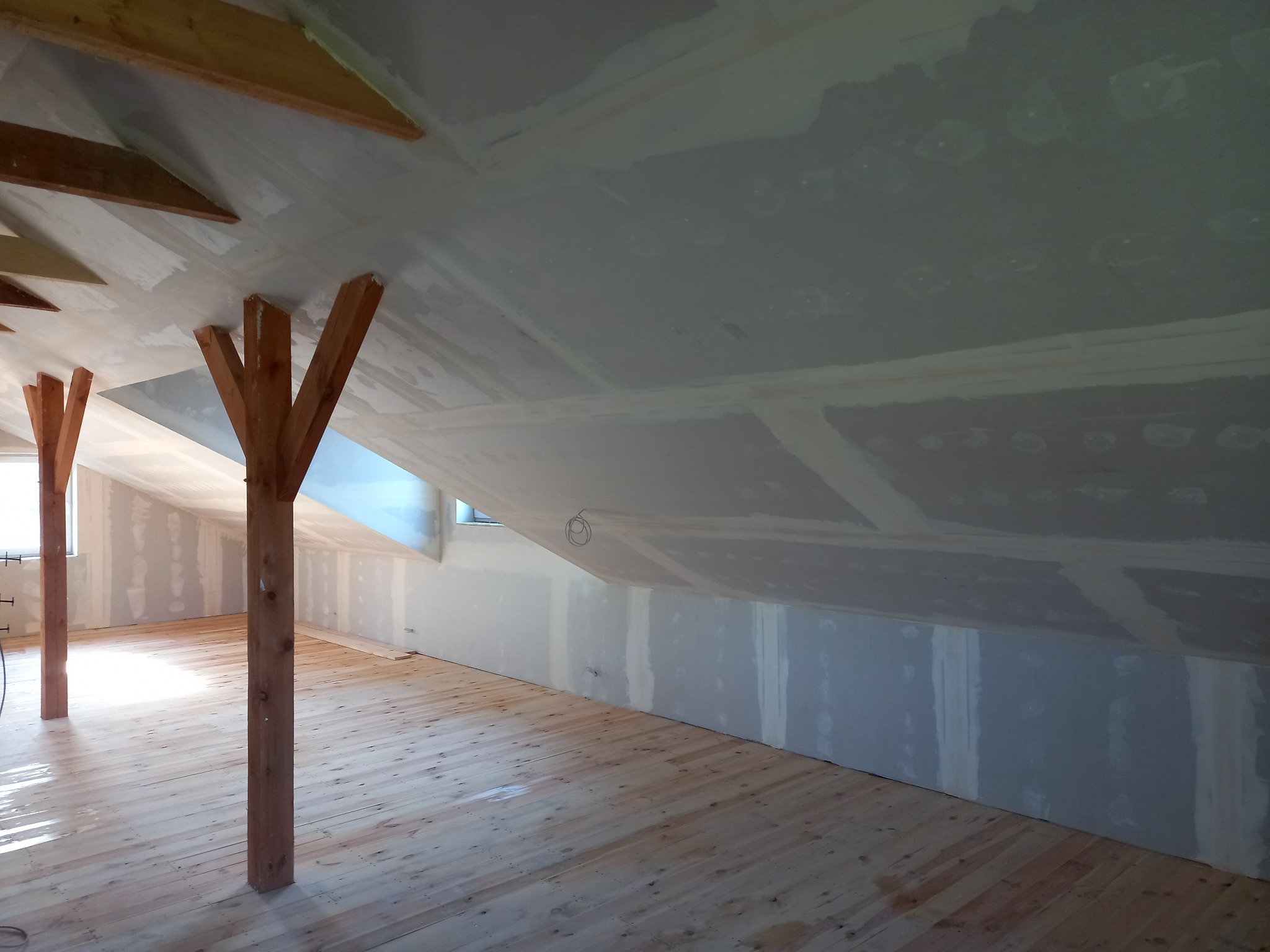 Poddasze po montażu płyt karton-gips na skosach, widoczne drewniane słupy konstrukcyjne i surowa drewniana podłoga, ściany przygotowane do dalszych prac wykończeniowych.