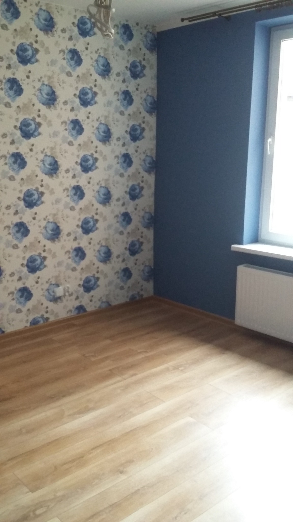 Pokój po remoncie z drewnopodobną podłogą, ścianą w kwiaty i granatową ścianą z grzejnikiem pod oknem.