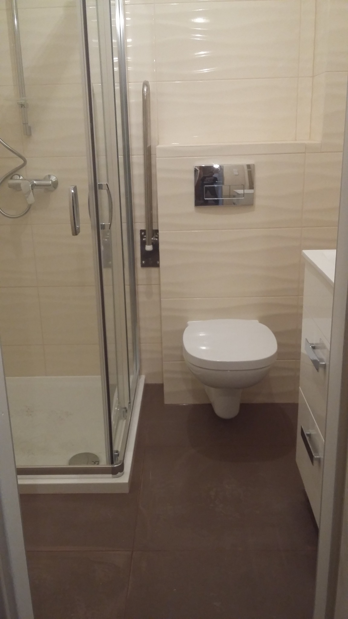 Nowoczesna łazienka z kabiną prysznicową, podwieszaną toaletą i brązowymi płytkami podłogowymi, wykończona w jasnych odcieniach beżu, z dodatkowym uchwytem dla osób z niepełnosprawnościami.