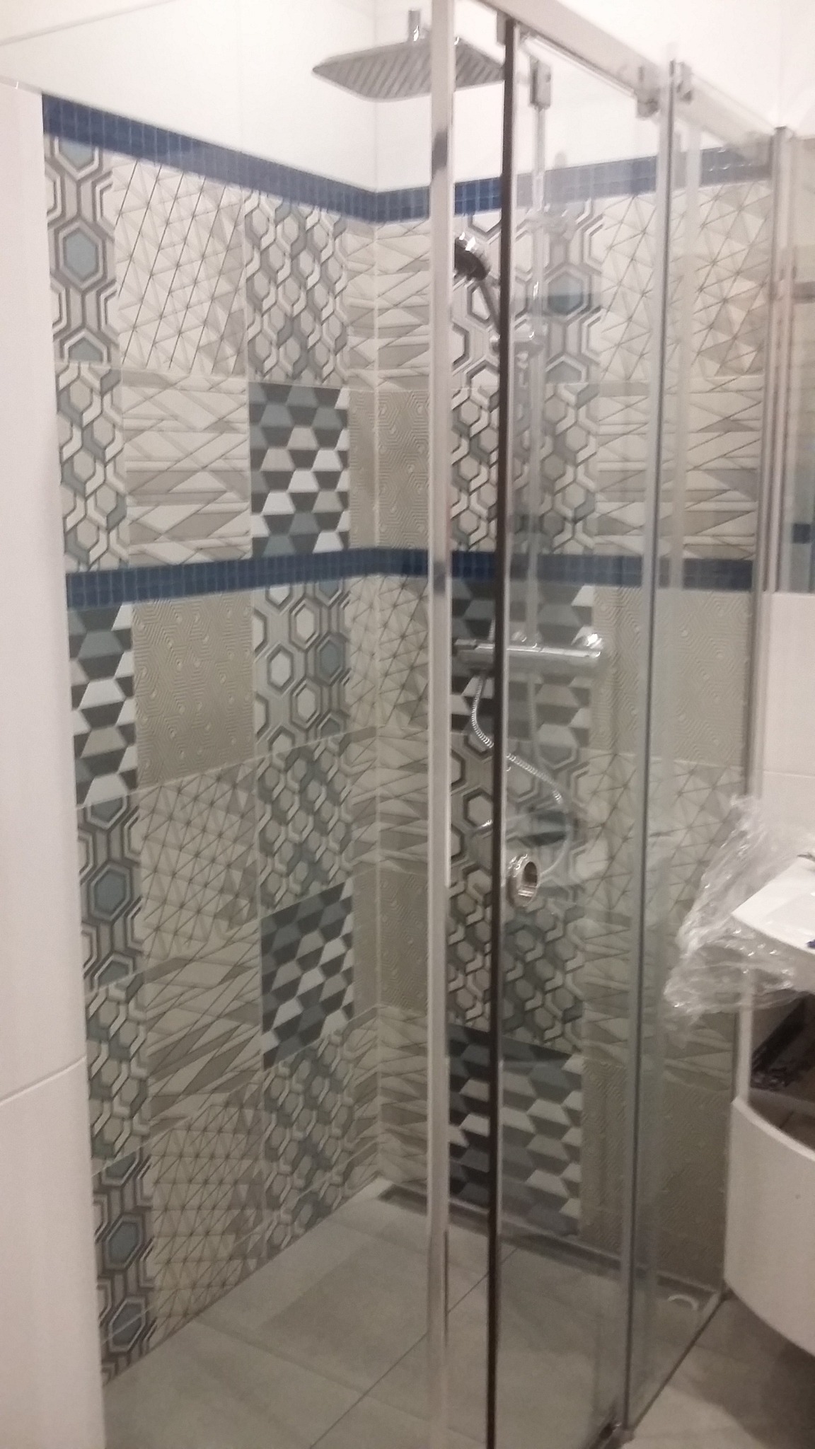 Nowoczesna kabina prysznicowa z geometrycznymi płytkami w odcieniach szarości i błękitu, wykończona chromowanymi elementami.