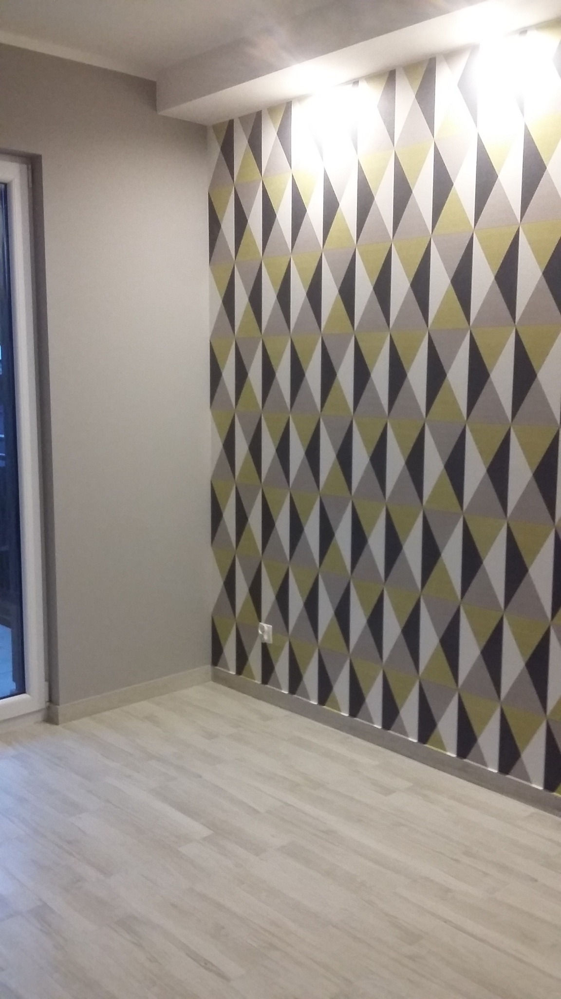 Pokój po remoncie: ściana z geometryczną tapetą w odcieniach szarości, bieli i żółci, jasna podłoga imitująca drewno, widoczne gniazdko elektryczne przy podłodze.