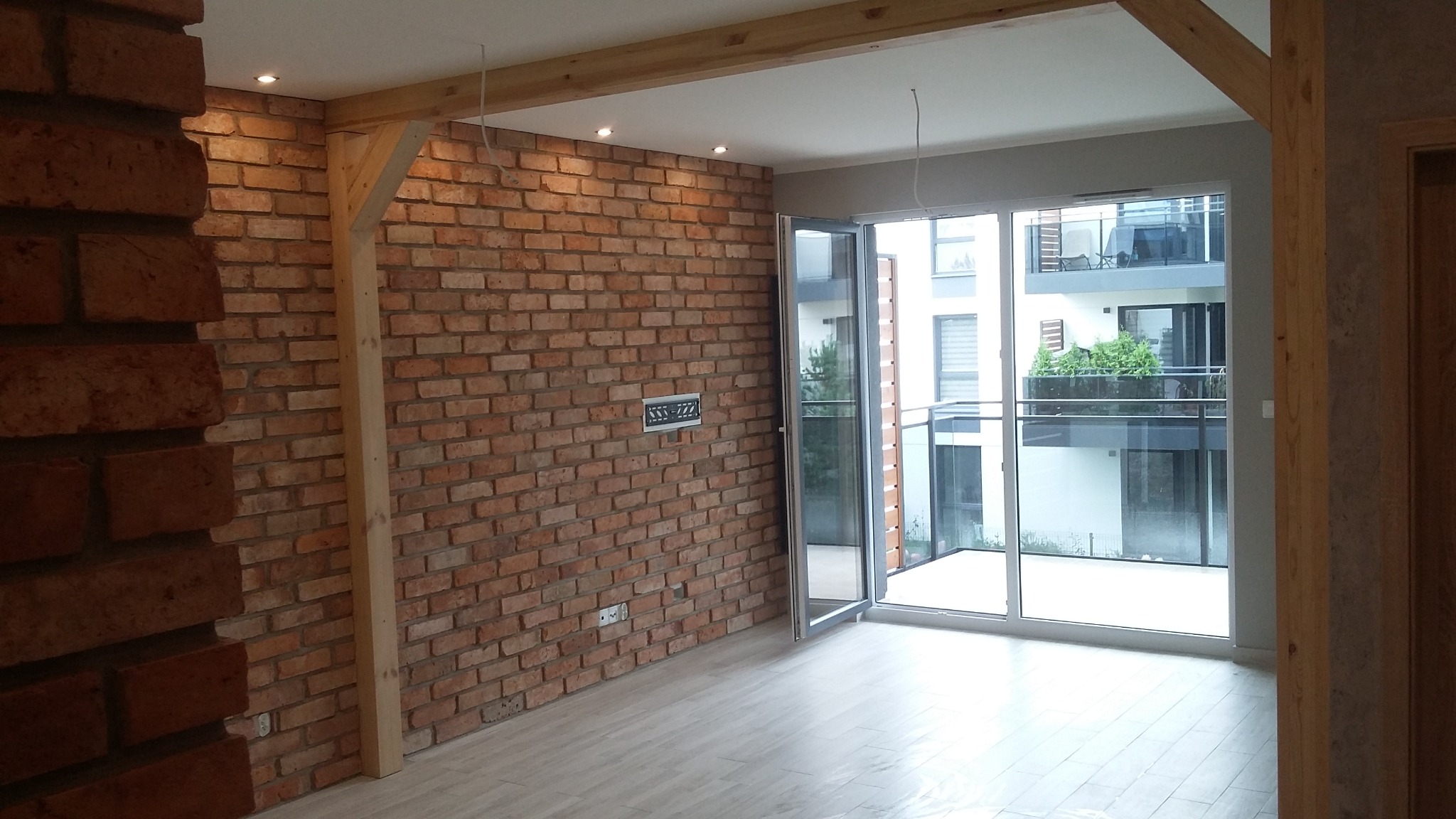 Wnętrze z ceglaną ścianą, drewnianymi belkami stropowymi i dużym oknem balkonowym z widokiem na nowoczesny budynek z balkonami.
