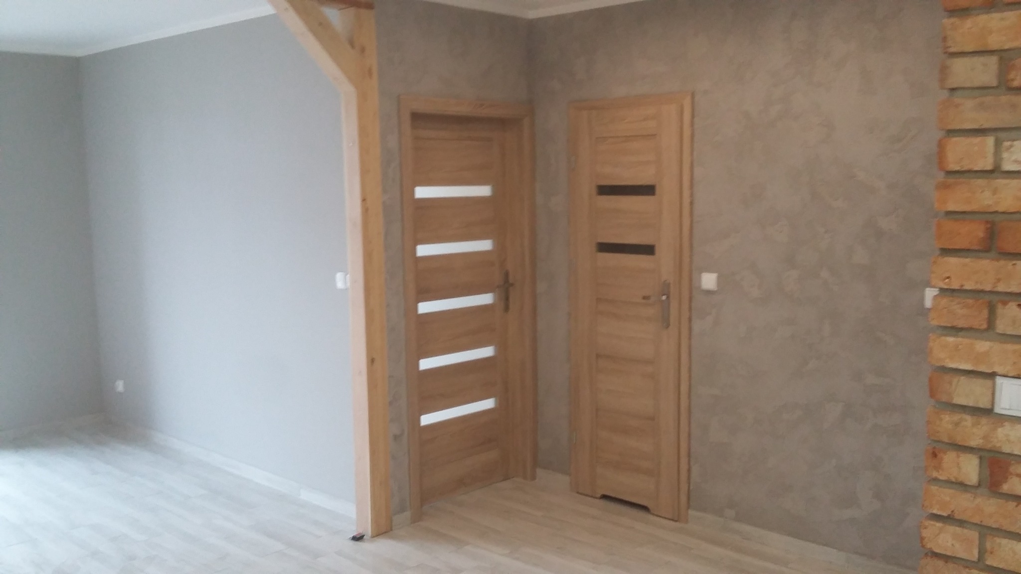 Dwie drewniane drzwi wewnętrzne z przeszkleniami, jedna z czterema poziomymi paskami, druga z jednym poziomym paskiem, zamontowane w pokoju po remoncie w Toruniu, z szarą ścianą i fragmentem ściany...