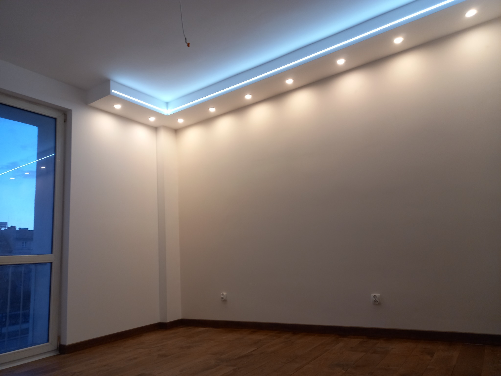 Pusty pokój z drewnianą podłogą, białymi ścianami i podwieszanym sufitem z oświetleniem LED oraz punktowymi lampami, widok na budynki za oknem.