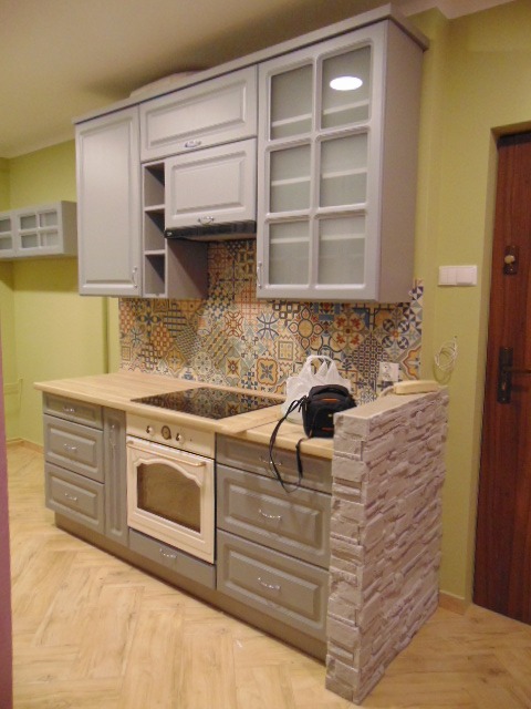 Szara zabudowa kuchenna z drewnianym blatem, wzorzystą mozaiką nad blatem i dekoracyjnym panelem imitującym kamień, widoczna kuchenka elektryczna i piekarnik w stylu retro.