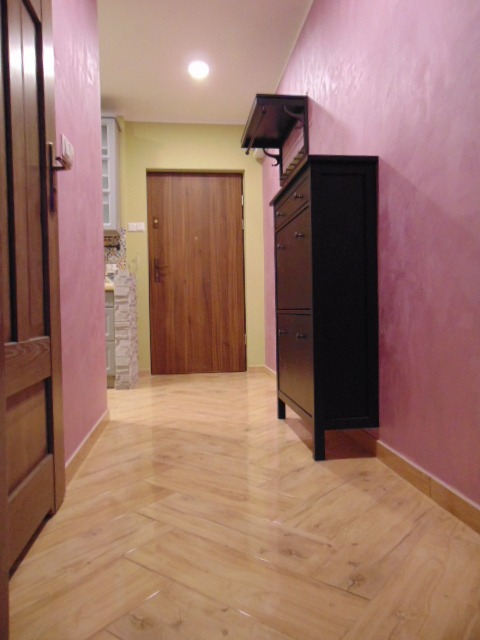 Przedpokój z podłogą ułożoną w jodełkę, ściany pomalowane na różowo z widoczną fakturą, ciemne meble do przechowywania i drzwi wejściowe.