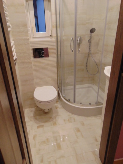 Jasna łazienka po remoncie: kabina prysznicowa z hydromasażem, podwieszana toaleta z chromowanym przyciskiem spłukiwania, kremowe płytki podłogowe i ścienne.