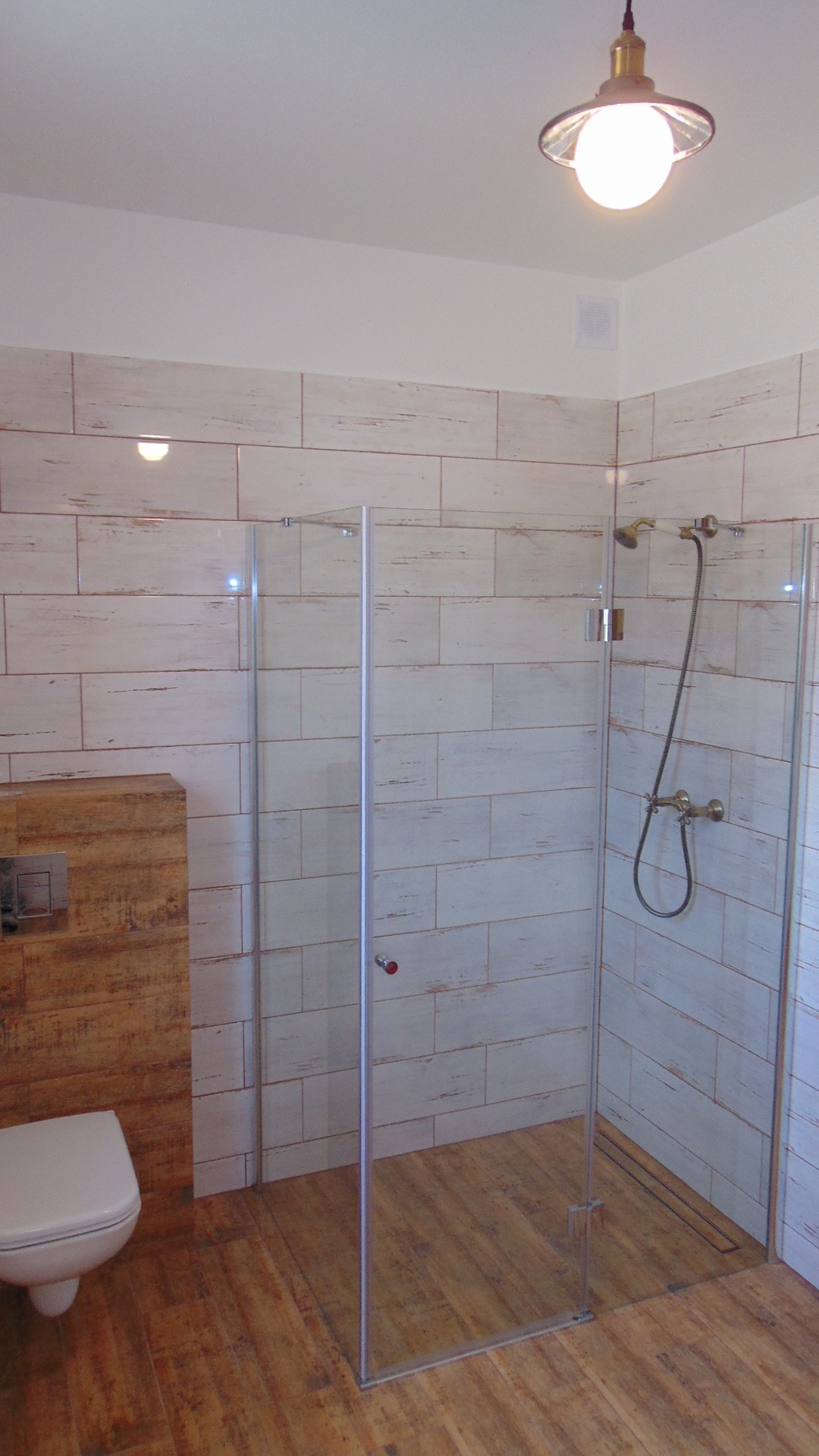 Nowoczesna kabina prysznicowa ze szklanymi ściankami, drewnopodobne płytki ścienne i podłogowe, wisząca toaleta z zabudową, oświetlenie sufitowe w stylu industrialnym.