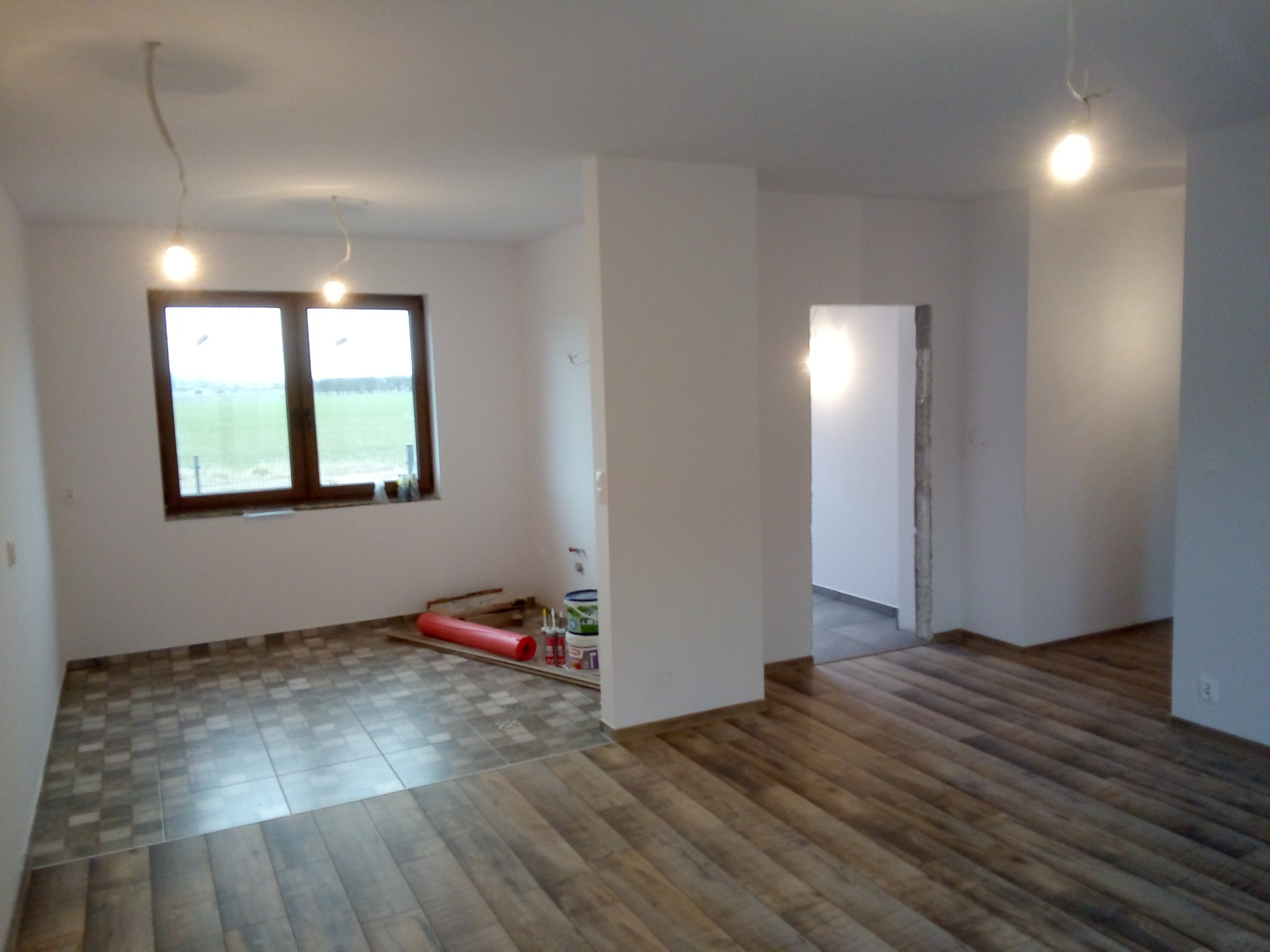 Wnętrze w trakcie wykańczania: połączenie płytek podłogowych imitujących drewno i szarej mozaiki, surowe ściany, widoczne materiały budowlane, okno z widokiem na zewnątrz, wiszące żarówki bez kloszy.