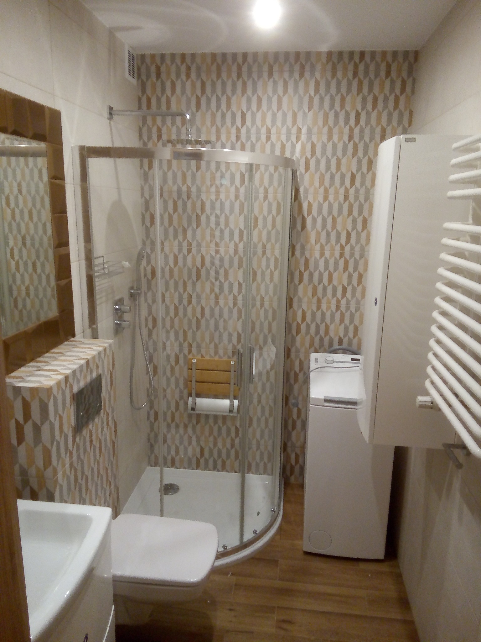 Nowoczesna łazienka z kabiną prysznicową typu walk-in, umywalką, toaletą i pralką, wykończona płytkami w geometryczny wzór w odcieniach brązu i beżu.