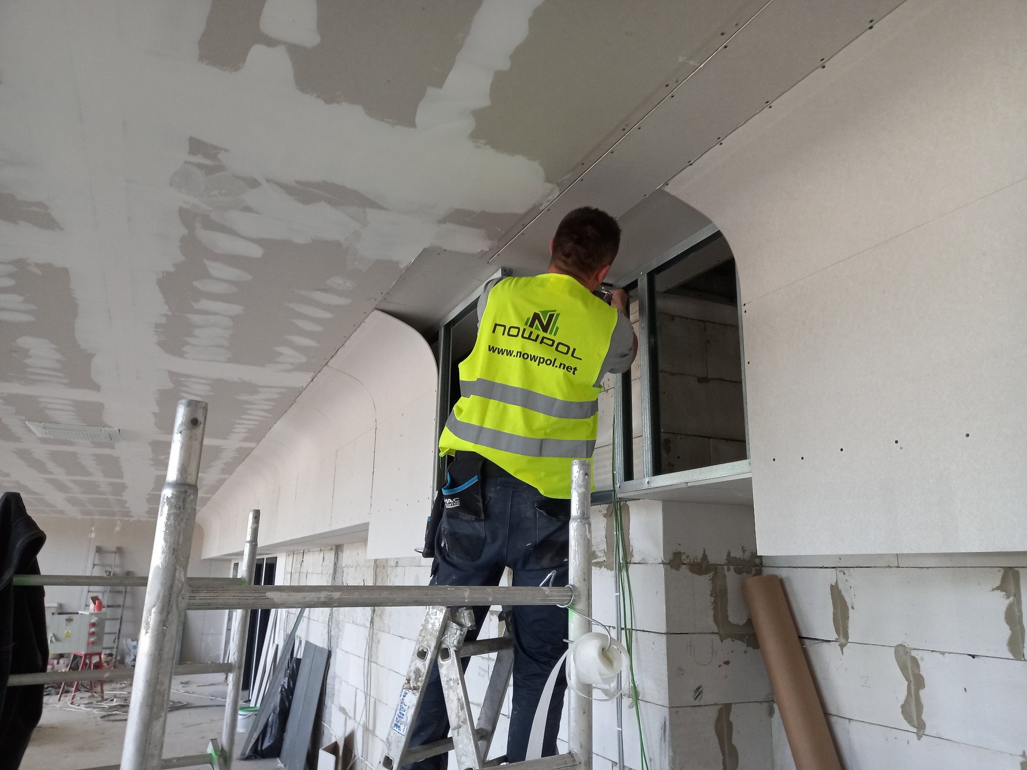 Pracownik w kamizelce odblaskowej montuje płytę kartonowo-gipsową na suficie, stojąc na aluminiowej drabinie; widoczne profile metalowe, szpachlowana powierzchnia sufitu i ściany z bloczków.