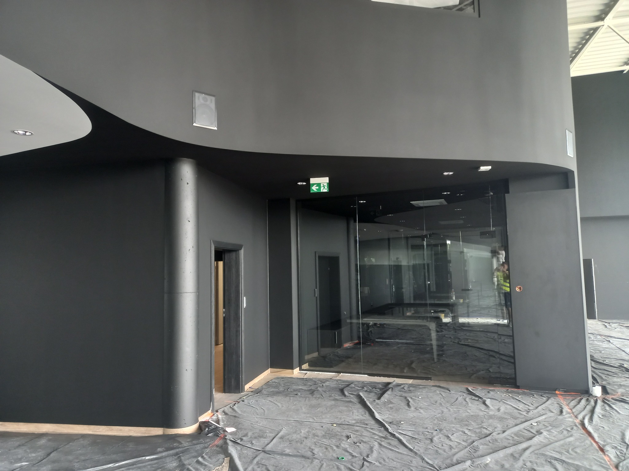 Nowoczesne wnętrze biurowe w trakcie malowania na kolor grafitowy, zabezpieczona podłoga folią ochronną, widoczne szklane ścianki działowe i elementy oświetlenia sufitowego.