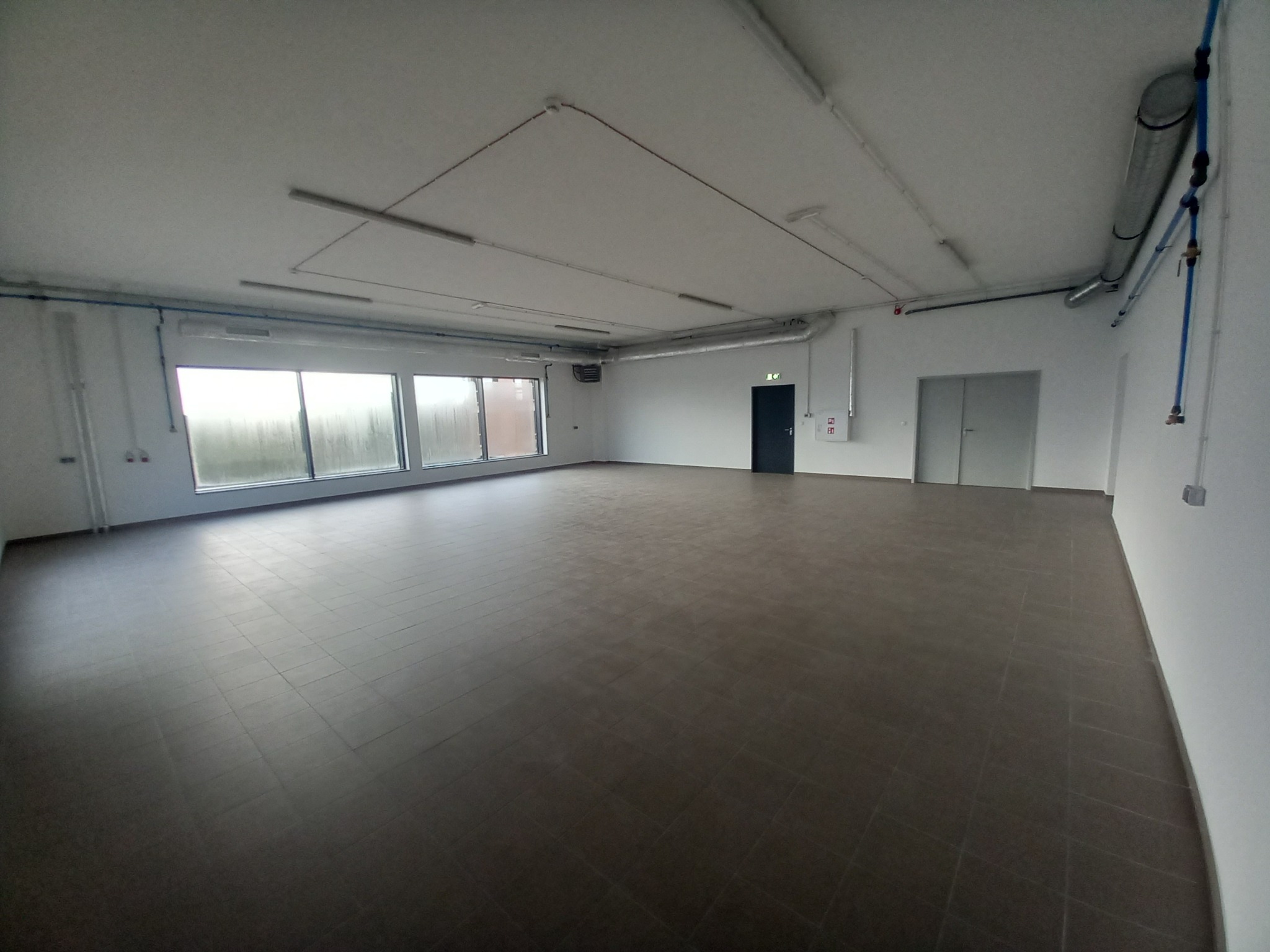 Duże, puste pomieszczenie z brązową podłogą z płytek, białymi ścianami, rurami pod sufitem i oknami z widocznymi kroplami deszczu.