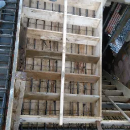 Szczegółowe ujęcie szalunku drewnianego z widocznym zbrojeniem stalowym przygotowanego do wylania betonu na schody, z fragmentem pompy do betonu w górnej części kadru.