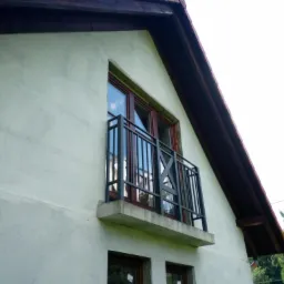 Balkon z czarną, metalową balustradą zamontowany na betonowej płycie, widok z dołu na elewację domu z oknami w brązowych ramach.