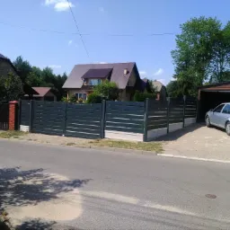 Nowoczesne, poziome ogrodzenie panelowe w kolorze grafitowym z betonową podmurówką, oddzielające posesję od drogi, z widocznym domem i garażem w tle.