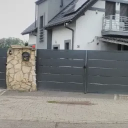 Nowoczesna, pozioma, grafitowa brama wjazdowa z kamiennymi słupkami i skrzynką na listy, na tle domu z balkonem i panelami słonecznymi.