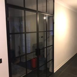 Usługi spawalnicze Roman Migdalek - Dwuskrzydłowe, przeszklone drzwi w stylu industrialnym z czarnymi szprosami, oddzielające korytarz od wnętrza domu, widoczne czarne płytki podłogowe i biała ściana z listwą przypodłogową.